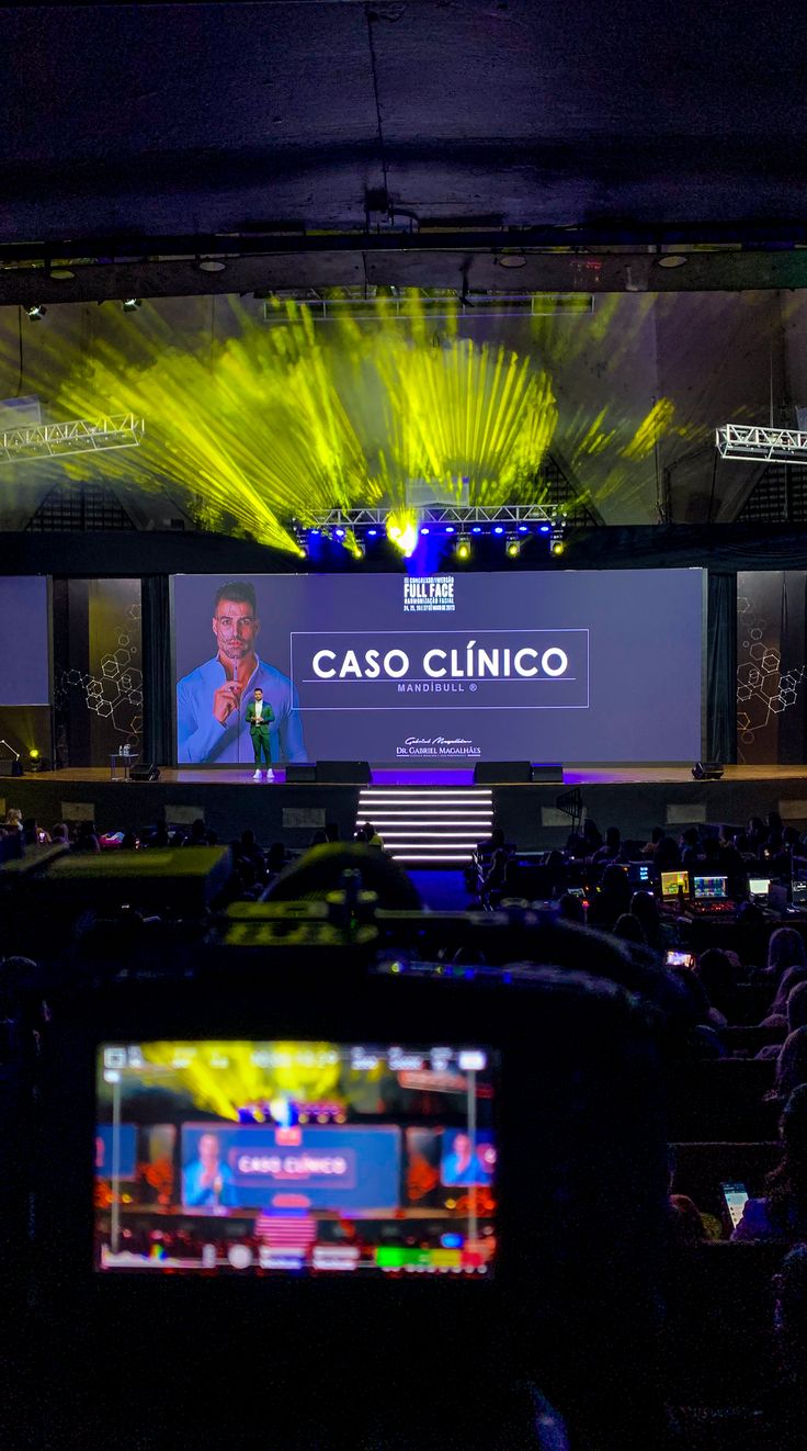 Palco com projeção, em evento corporativo, visto de dentro da house de áudio visual com destaque para luzes verdes no teto 